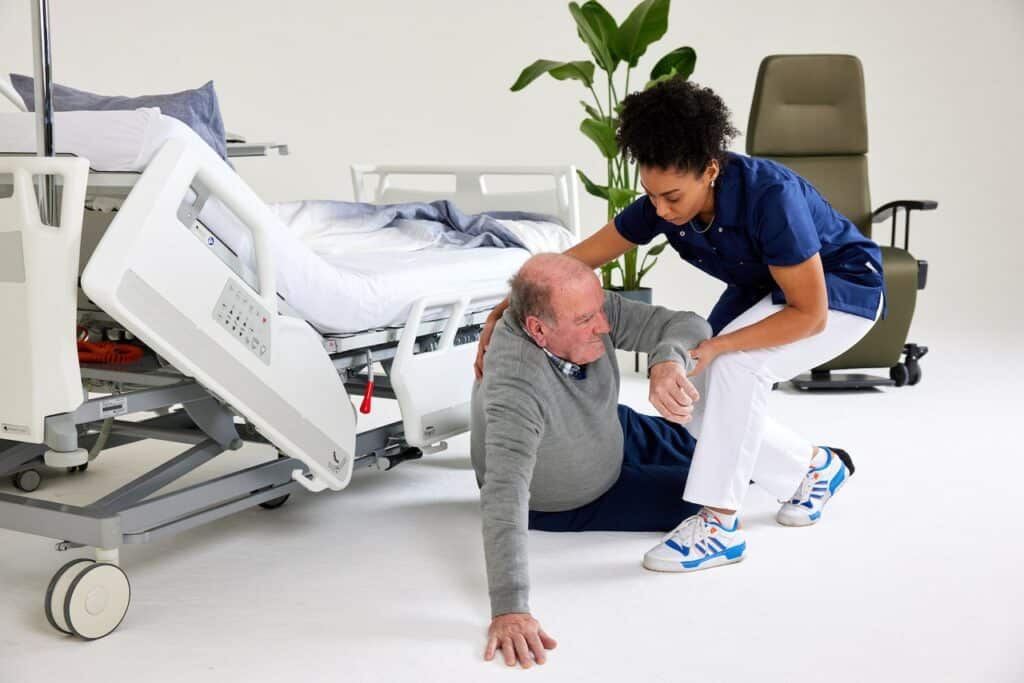 Meneer is naast zijn bed gevallen. Een verpleegkundige helpt hem overeind.